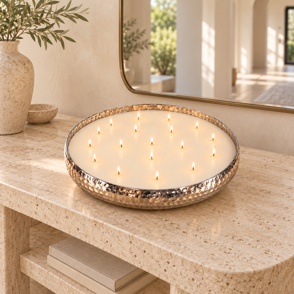 A large round wax candle in a hammered metal bowl, lit with multiple small flames, set on stone piece, in a bright and white hallway.