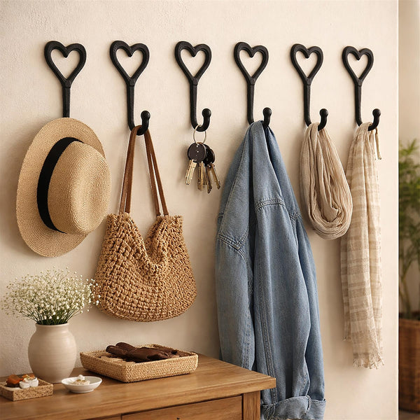 Six black heart-shaped wall hooks displayed on a beige wall above a wooden console table, styled with a straw hat, woven shoulder bag, keys, denim jacket, cream scarf, and plaid scarf.
