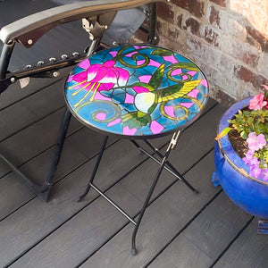 Colorful stained-glass table on a wooden deck with a chair and potted plant.