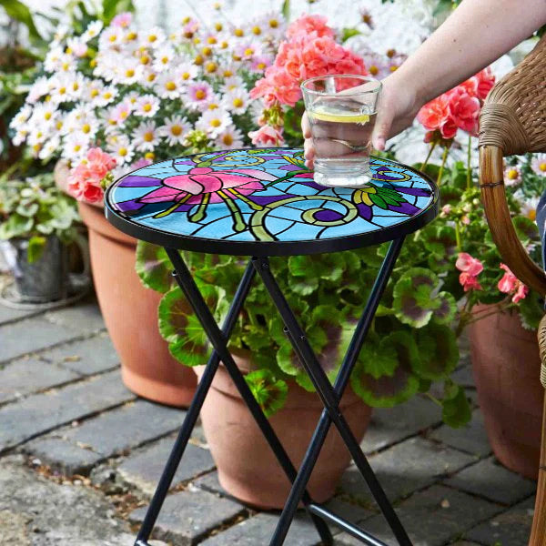 Colorful mosaic table in a garden setting with a person holding a glass.