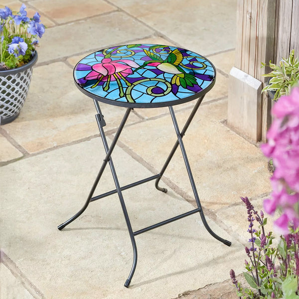 Colorful stained-glass table on a stone patio with flowers.