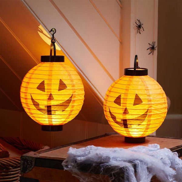 Two glowing orange paper lanterns with jack-o'-lantern faces hanging in a home setting, with Halloween decorations including fake cobwebs and plastic spiders on the wall.
