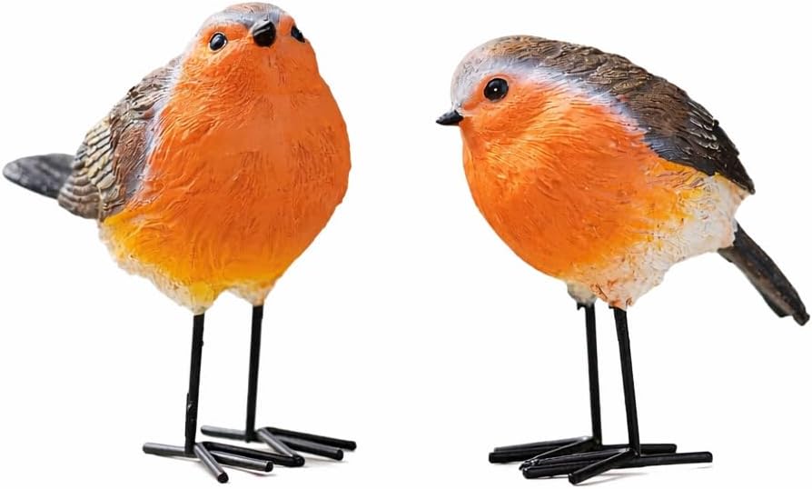 Two decorative bird figurines on a wooden surface with flowers and a watering can in the background.