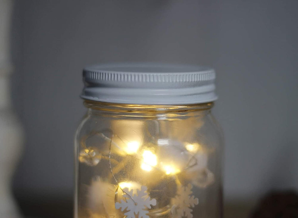 A glass jar with a silver lid, decorated with white LED lights and snowflake patterns, providing a warm glow.