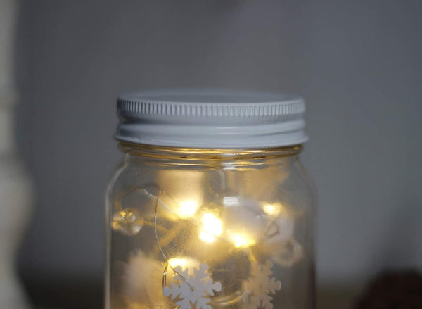 Mason jar with white lid containing string lights and snowflake decorations on a dark background