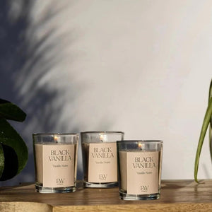 Three 'Black Vanilla' candles on a wooden surface with a plant in the background