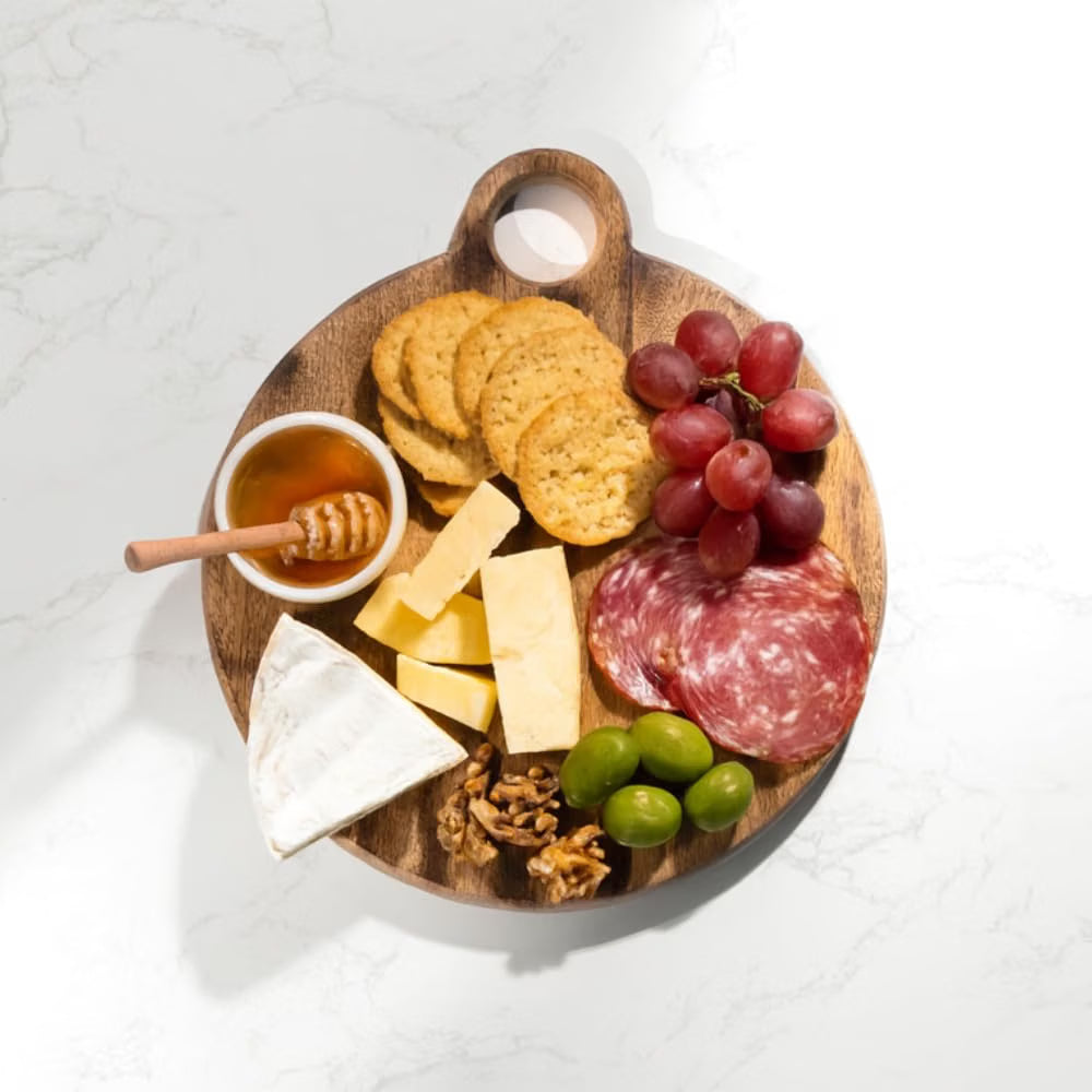 A round wooden charcuterie board with a handle, styled with crackers, grapes, cheese, salami, olives, walnuts, and a small cup of honey, photographed from above on a white marble surface.