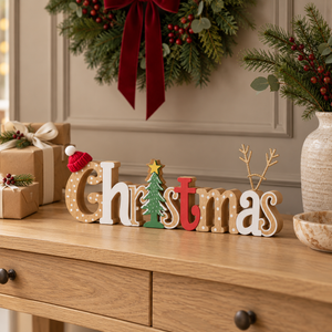 Wooden decorative letters spelling Christmas arranged on a wooden console table, with a green wreath and holiday decorations in the background.