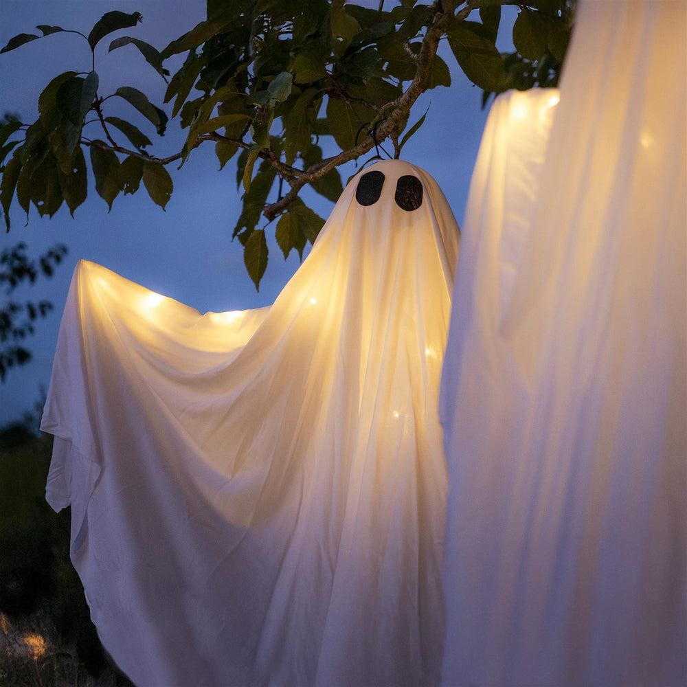 Decorative ghost made of white fabric with lights, hanging from a tree at night.