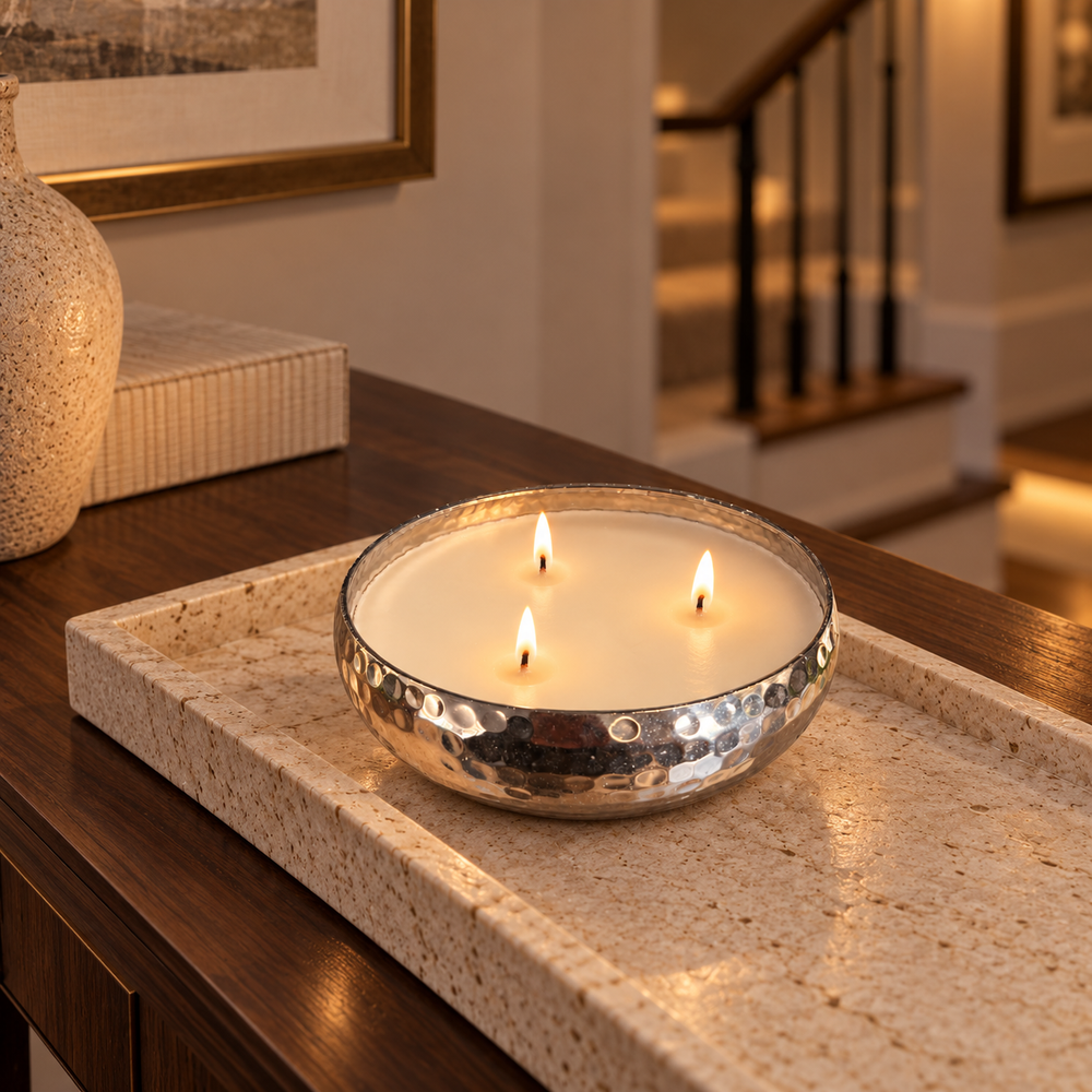 A three-wick soy candle in a hammered silver metal bowl sits on a beige stone tray on a dark wood console table in a warmly lit home interior.