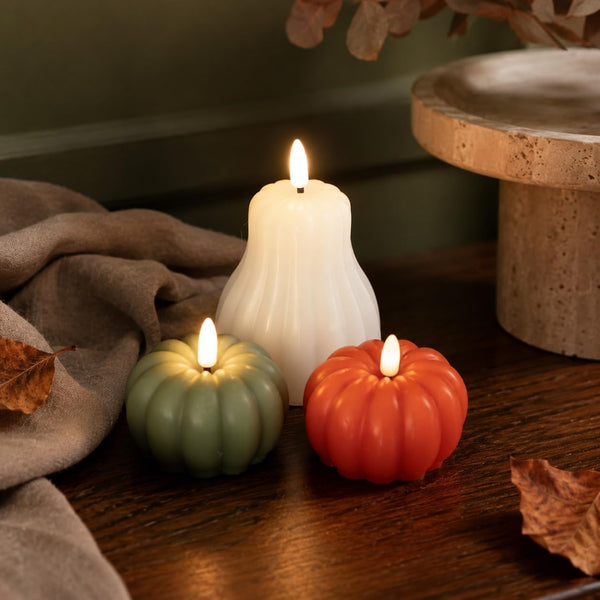 Three flameless LED pillar candles shaped like a white bell candle and two small pumpkin-shaped candles in sage green and terracotta orange, styled on a dark wooden table with autumn leaves and a neutral fabric.