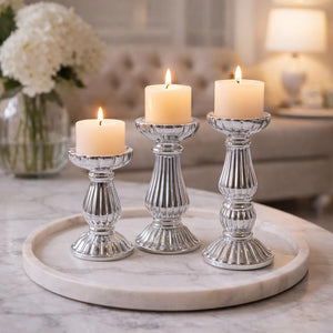 Three silver fluted glass candlestick holders with lit cream pillar candles, arranged on a round white marble tray in a softly blurred living room setting.