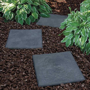 Three dark gray stepping stones on a mulched garden bed with green plants.