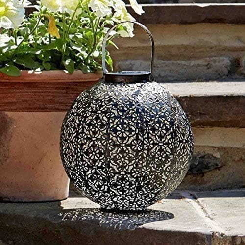 A round black metal lantern with an intricate laser-cut geometric pattern and a top handle, placed on stone steps next to a potted plant.