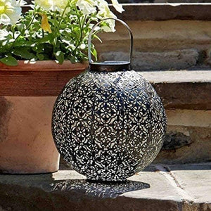 A round black metal lantern with an intricate laser-cut geometric pattern and a top handle, placed on stone steps next to a potted plant.
