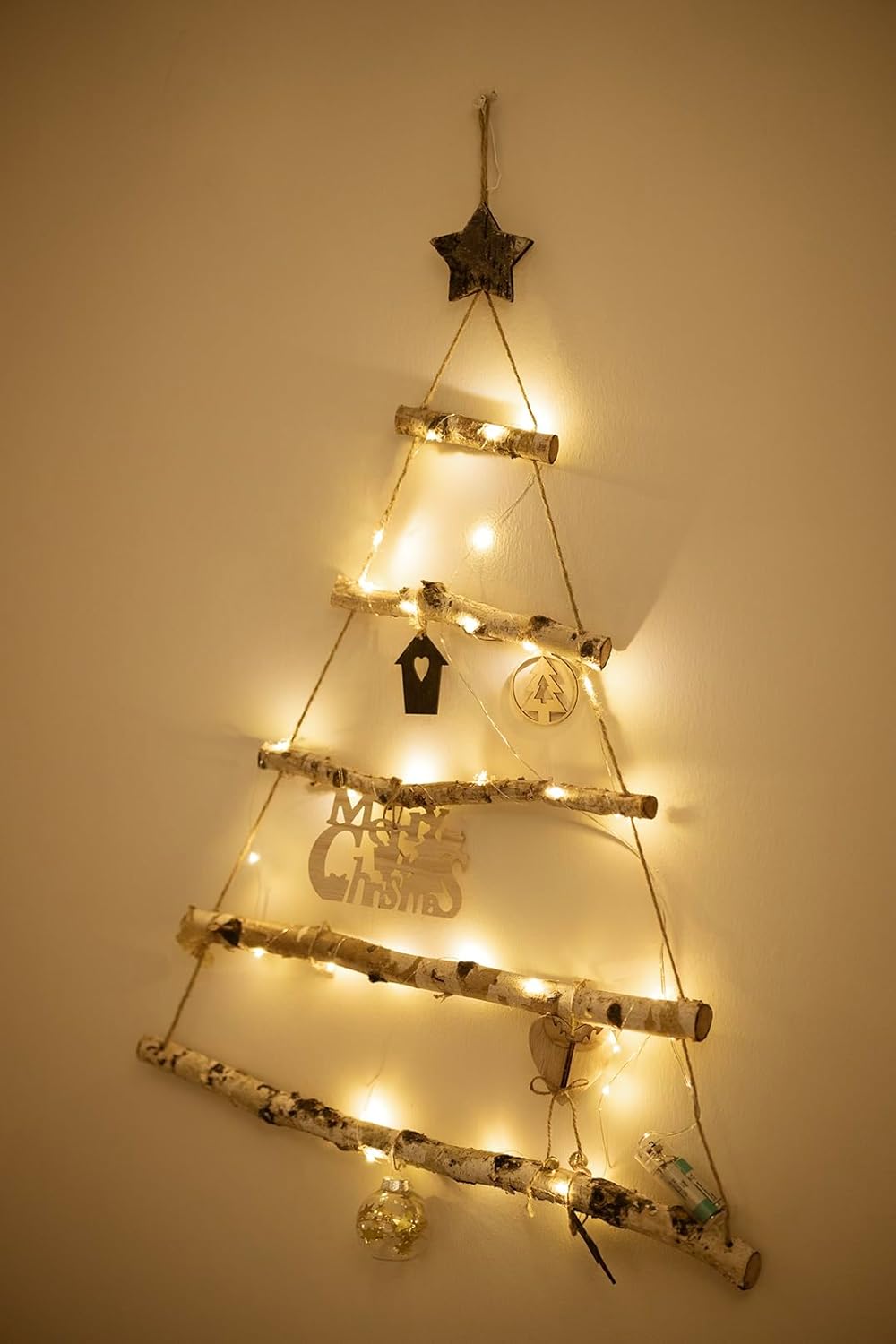 Decorative wall-mounted Christmas tree made of branches with lights and small ornaments on a plain wall.