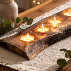 A rustic live-edge wooden candle holder with four lit tealight candles, styled on a textured cream table runner with eucalyptus leaves and a neutral ceramic vase in the background.