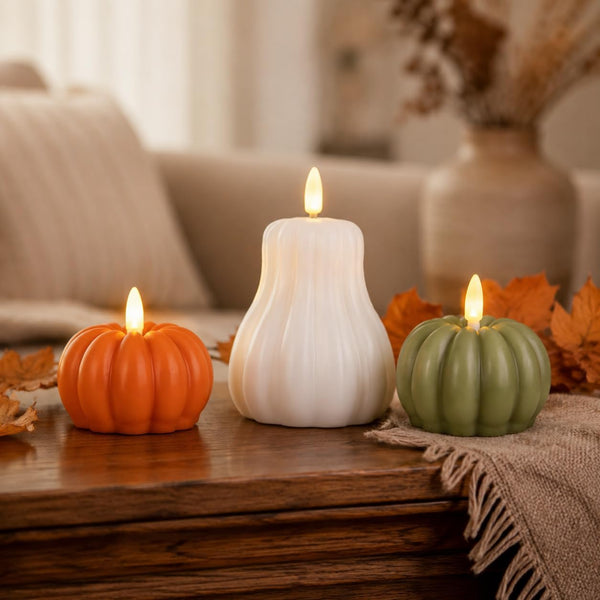 Three decorative pumpkin-shaped candles in orange, white, and sage green are arranged on a wooden table in a cozy living room setting with autumn leaves and a throw blanket.
