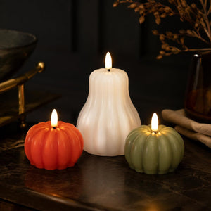 Three flameless LED pumpkin candles in orange, white, and sage green, arranged on a dark tabletop with warm lights glowing from the wicks.