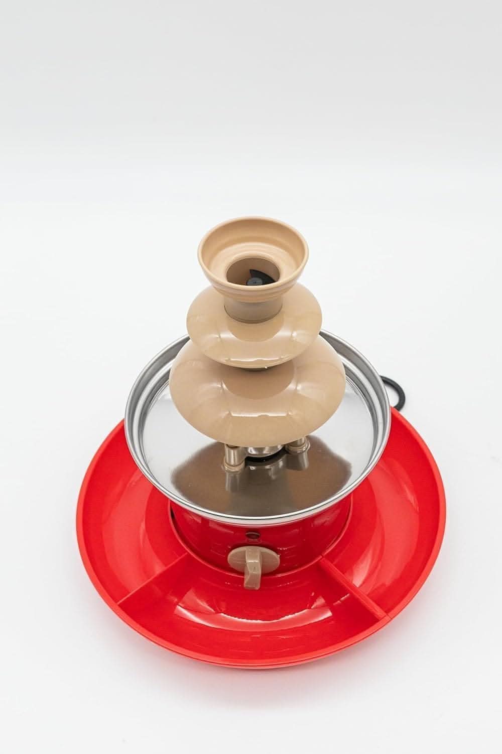 A 3 tier chocolate fountain on a wooden board with a red tray at the bottom. The fountain is on a kitchen counter with chocolate pieces scattered around and a plant in the background.