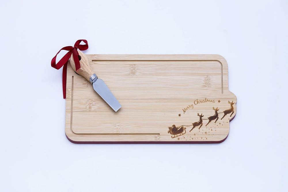 A Christmas-themed wooden cheese board with a Santa sleigh design, displayed on a table with cheese and crackers, a knife, and holiday decorations in the background.