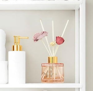 White dispenser bottle with gold pump and pink diffuser sticks with flowers on a white shelf.