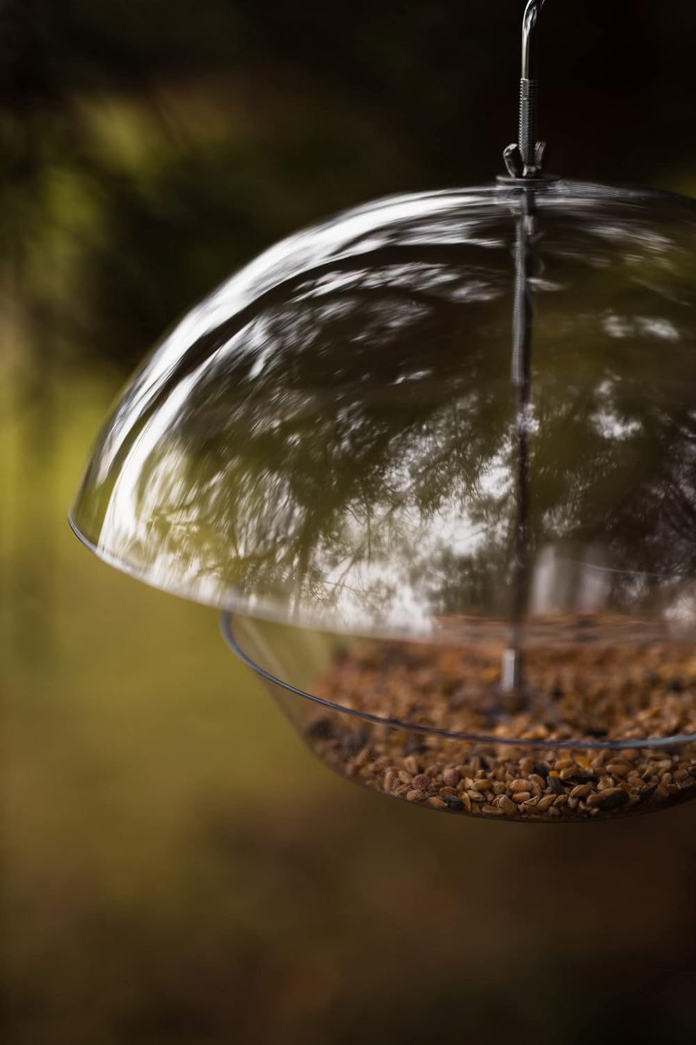A clear glass bird feeder bowl filled with brown birdseed, suspended by a metal wire in an outdoor setting with a blurred green and brown background.