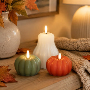 Three flameless LED pillar candles shaped like a white bell candle and two small pumpkin candles in sage green and orange, styled on a wooden table with autumn leaves, a knitted blanket, and a vase.