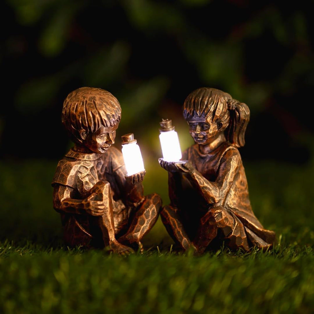 Two bronze-finished garden statues of children sitting on grass at night, each holding a glowing lantern. The background is a dark, blurred outdoor setting.