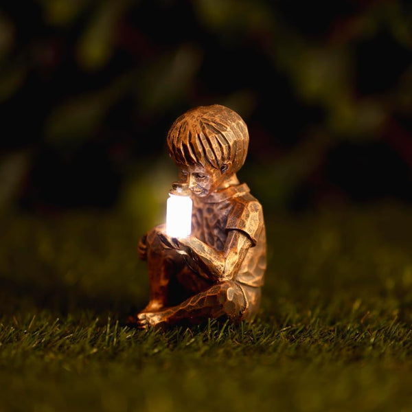 A small bronze-finished garden gnome figurine sitting on grass at night, holding a glowing white candle-like light. The background is dark and blurred foliage.