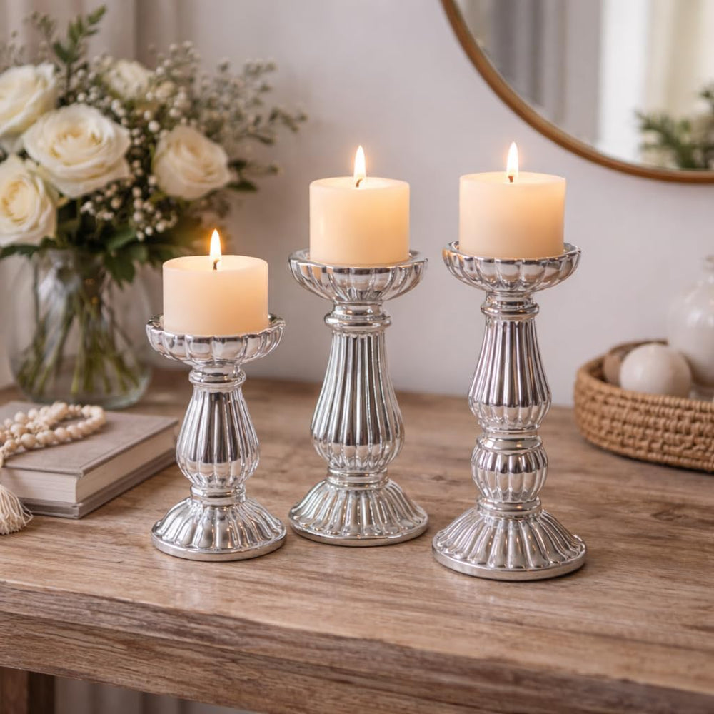 Three silver candlestick holders with lit cream pillar candles, styled on a wooden table with a vase of white roses and a mirror in the background.