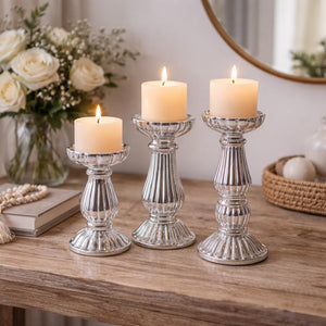 Three silver candlestick holders with lit cream pillar candles, styled on a wooden table with a vase of white roses and a mirror in the background.