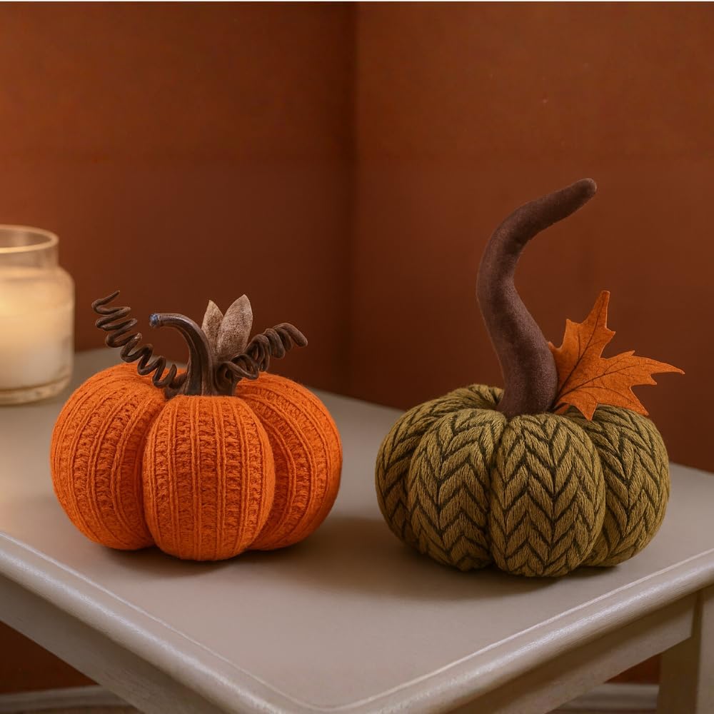 Two knitted pumpkins, one orange and one green, on a table with a candle in the background.