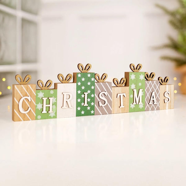 Wooden Christmas letter sign spelling CHRISTMAS, styled as wrapped gift boxes in green, beige, white, and brown patterns, with small wooden bows on top, displayed on a white surface.