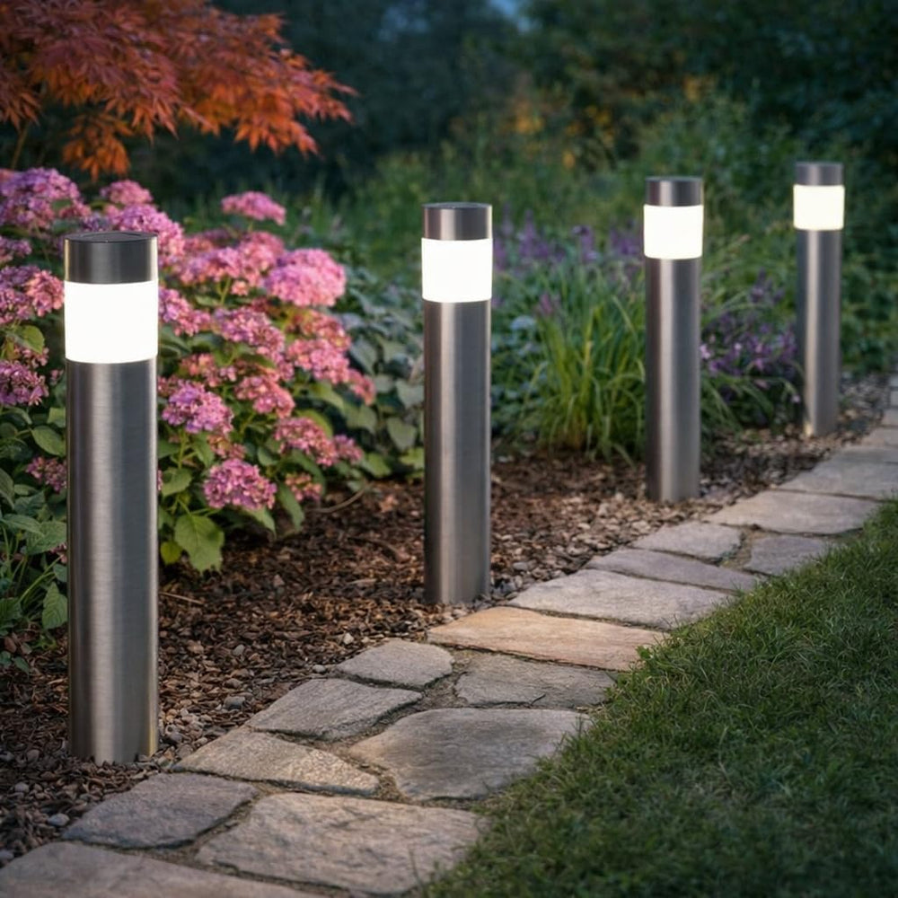 Four modern outdoor bollard pathway lights with brushed metal posts and glowing white cylindrical shades, installed along a stone garden path in a landscaped yard with pink flowers and green grass.