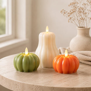 Three flameless LED pumpkin-shaped candles in green, white, and orange, styled on a round stone table with a neutral background and a vase of dried flowers.