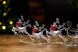 Decorative metal sleigh with reindeer and a small house on a wooden surface, blurred lights in the background.