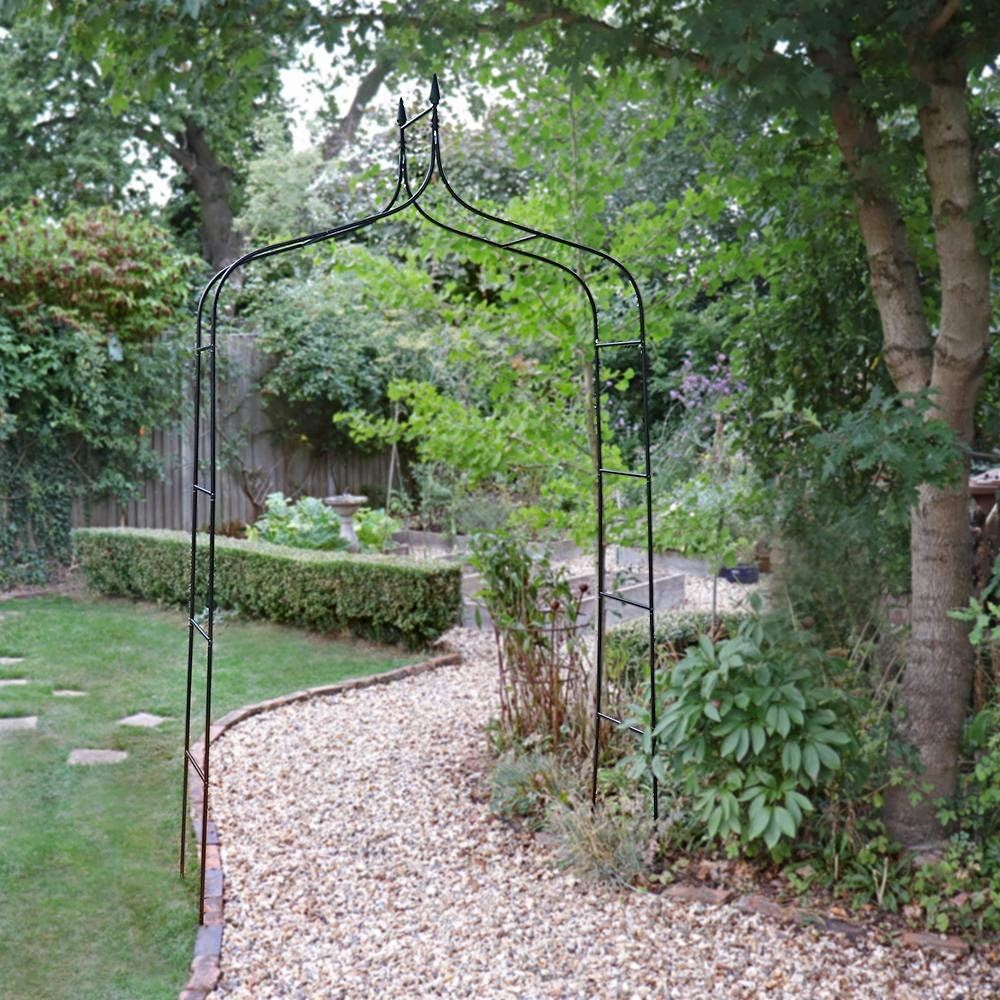 A green metal garden arch standing on a gravel path with greenery in the background.