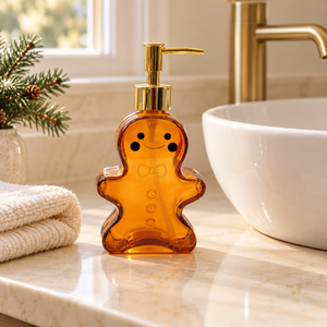A gingerbread-shaped amber glass soap dispenser with a gold pump and handle, styled on a stone tray next to a white sink and cream towel.