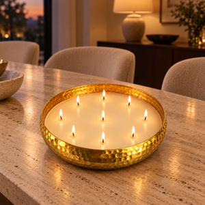 A hammered gold metal candle holder filled with multiple lit white candles, placed on a beige stone countertop in a warmly lit dining or living room setting.