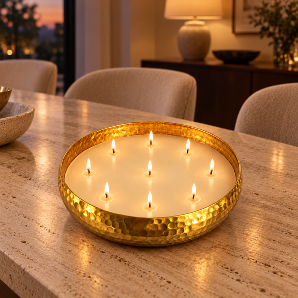 A hammered gold metal candle holder filled with multiple lit white candles, placed on a beige stone countertop in a warmly lit dining or living room setting.