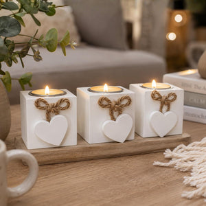 Three white square candle holders with heart decorations and twine bows, each holding a lit tea light candle, arranged on a wooden tray on a table in a cozy indoor setting.