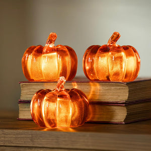 Three glowing orange glass pumpkins on stacked books with a warm glow.