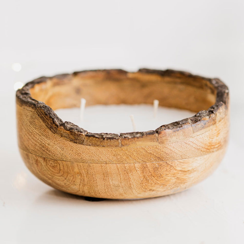A three-wick soy wax candle in a rustic, handcrafted wooden bowl with a natural bark edge, lit with three small flames on a white tabletop.