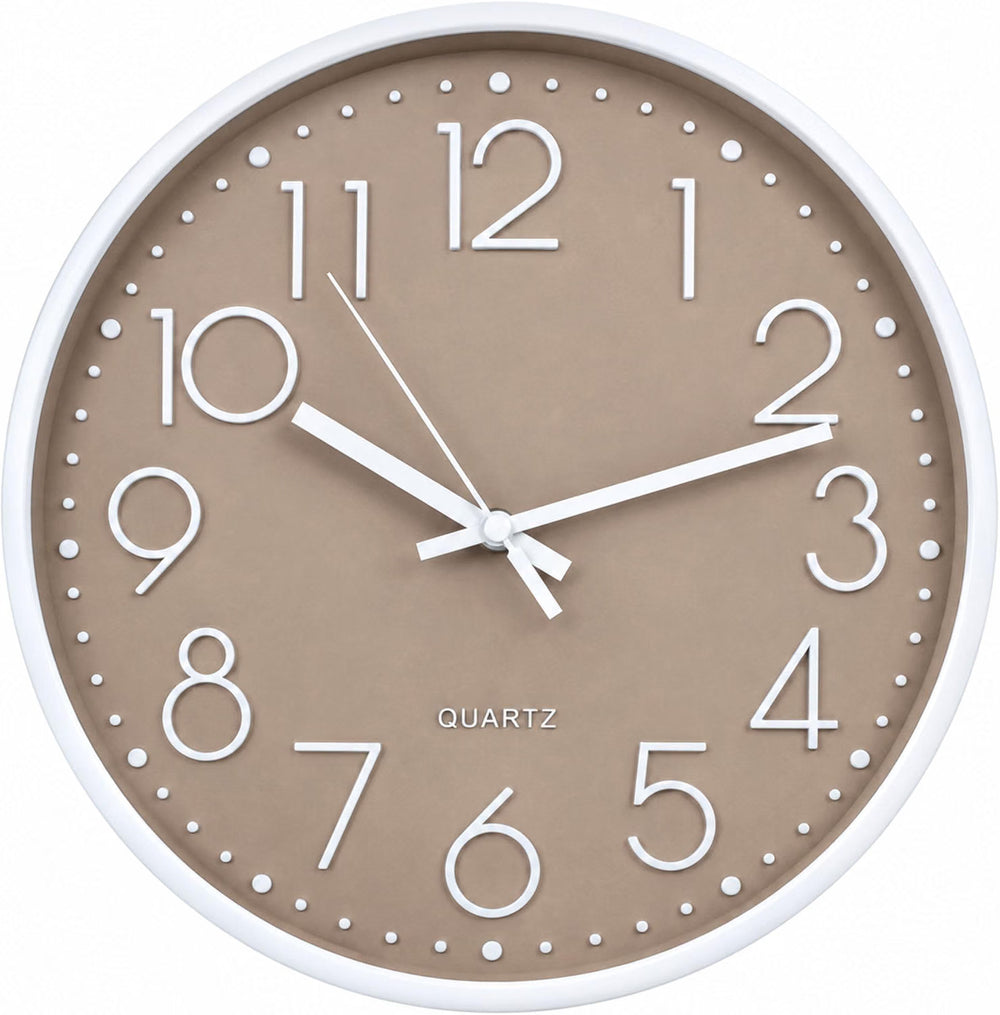 A round wall clock with a white frame and a warm taupe-beige face hangs on a white wall in a bright kitchen setting. The clock features white hands and black Arabic numerals.