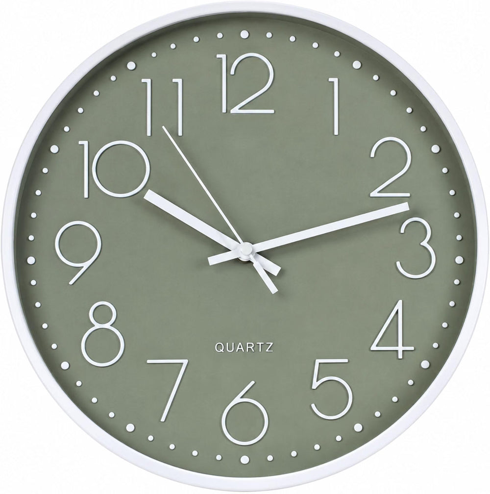 A round wall clock with a sage green face and white frame hangs on a light wall above a wooden countertop. The scene includes potted green plants, white canisters labeled TEA and COFFEE, and a bowl of green apples, creating a bright kitchen or cafe-like interior setting.