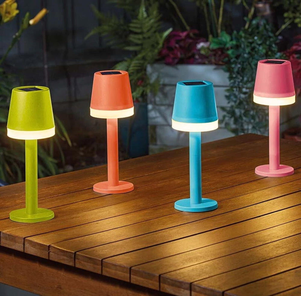 Four colorful outdoor lamps on a wooden surface with plants in the background