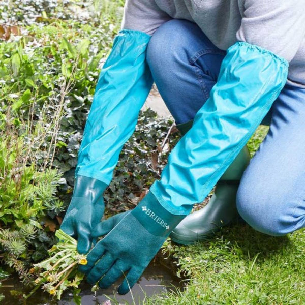 A person wearing blue waterproof gardening gloves up to the calf, crouching by plants.