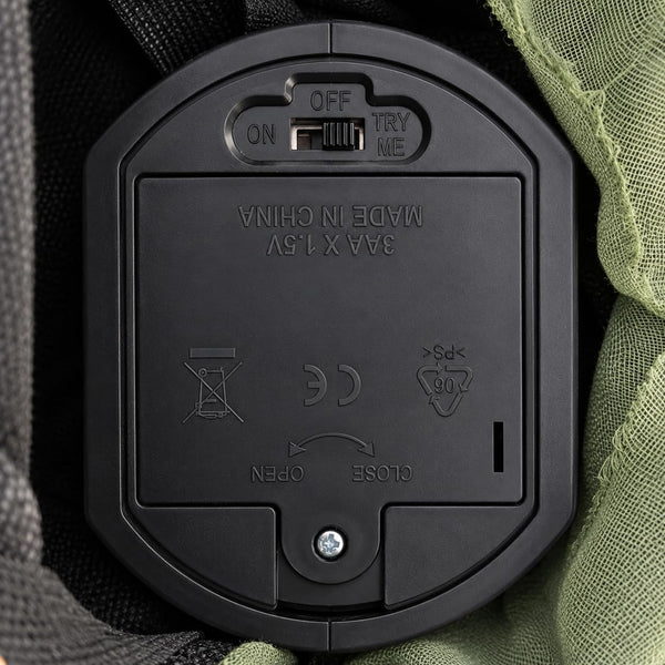 Close-up of a black plastic battery compartment cover with a switch labeled OFF/ON and TRY ME, showing embossed text 'MADE IN CHINA' and product symbols, set against dark fabric and olive green mesh.