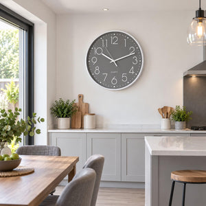 A round wall clock with a dark gray face and white numerals, mounted on a light gray wall in a modern kitchen setting.